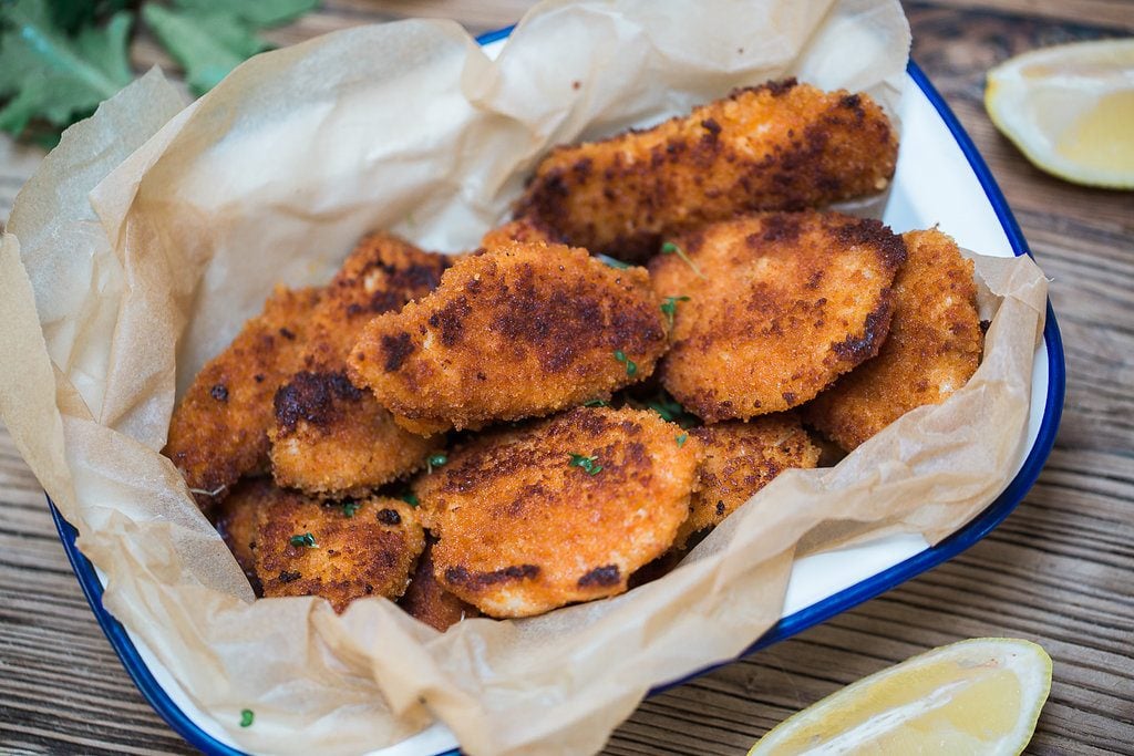 A basket with parchment paper full of homemade chicken nuggets, on a wood surface, surrounded by fresh lemon wedges. 