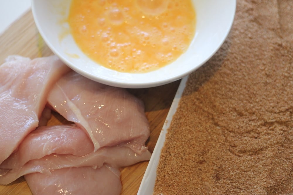 Raw chicken, a bowl of whisked eggs, and a shallow pan of gluten-free breading, sitting on a wooden cutting board. 