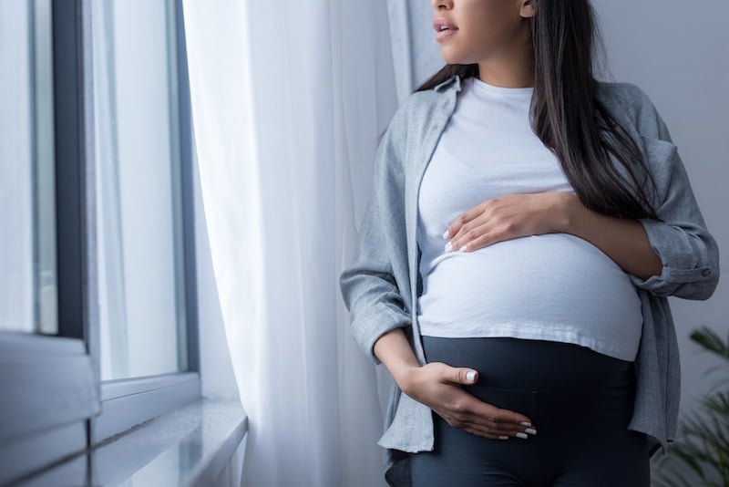 Pregnant woman standing by window in neutral clothing with hands on belly